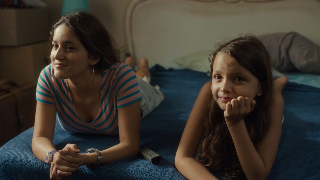 Two Latina girls lying on a blue bedspread are propping themselves up with their arms.