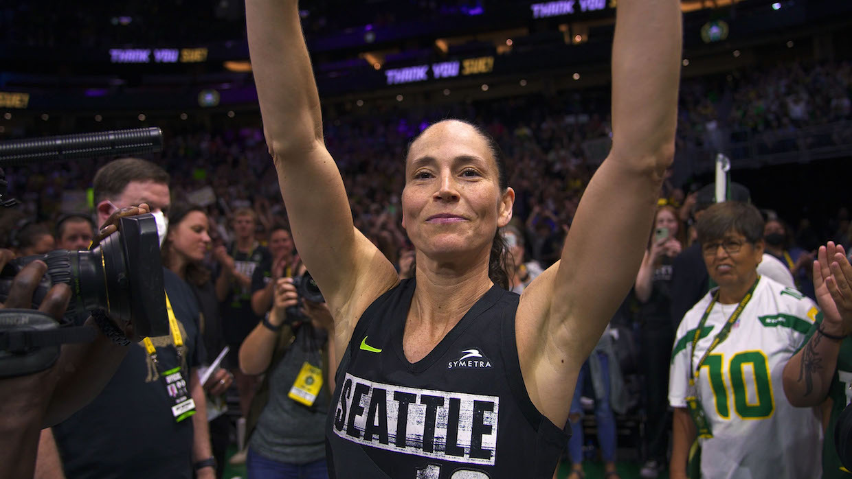 Basketball player Sue Bird raises her arms in victory and smiles at the camera.