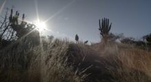 With the sun low and the sky, a distant figure are visible against a desert landscape.