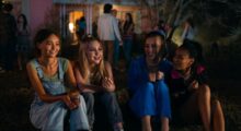 In the foreground, a group of adolescent girls smile. In the background, a group of adults are talking to one another.