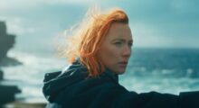 A young woman played by actress Saoirse Ronan with orange hair wearing a black hoodie standing against the sea