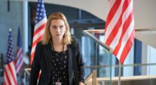 A young woman with blond hear wears a blazer and stands in a building lobby adorned with American flags.