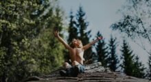 A blond, shirtless young boy sits in a forest, arms outstretched toward the sun.