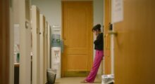 A woman in a black top and pink pants holds her face in despair in a deserted hallway.