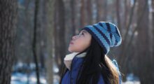 A young Japanese girl in a knit beanie stands staring upwards in a sunny forest during winter.