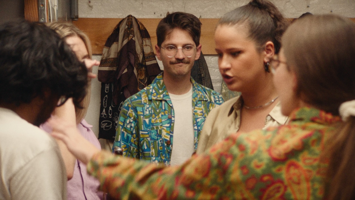 A white man with a mustache smirks at the back of a crowded room.
