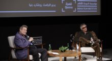 Two men sit on a stage separated by a small wooden table at a Q&A.