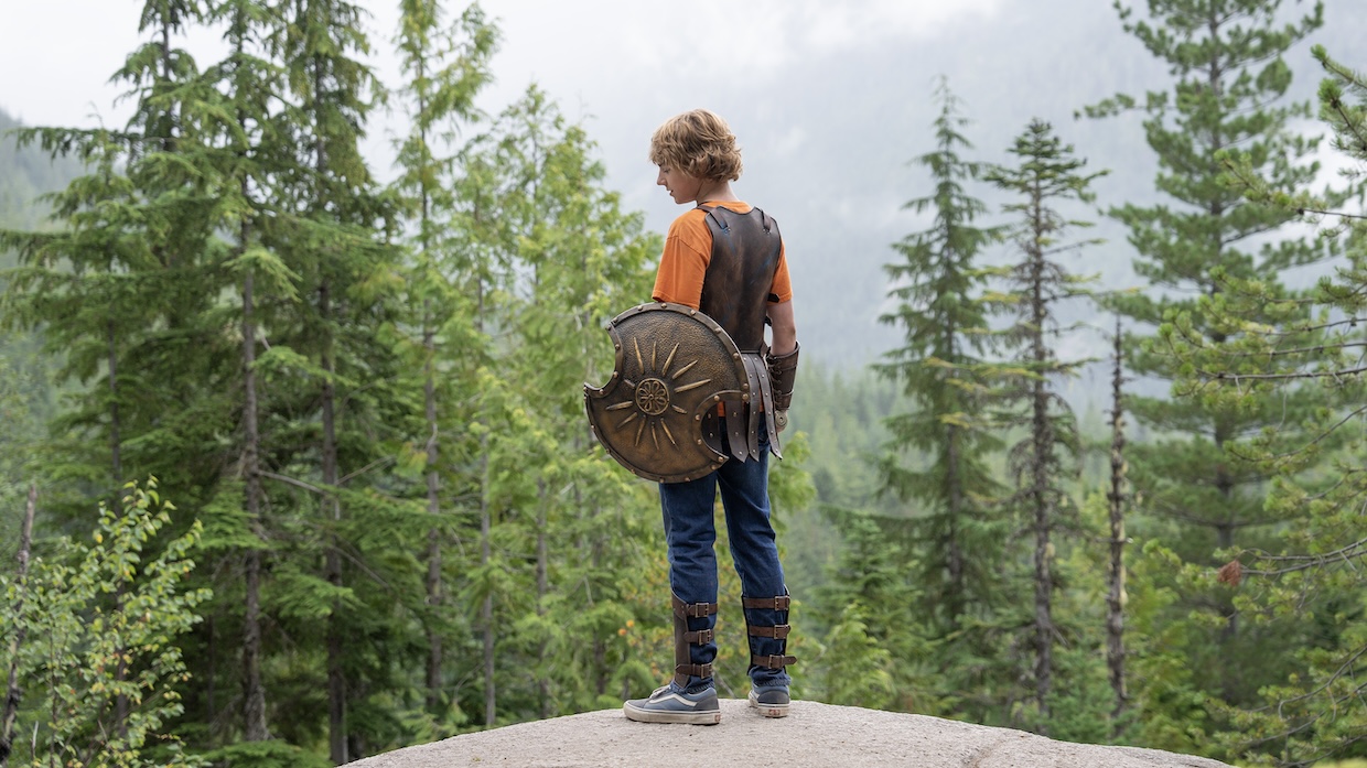 A young man holding a shield with a chipped section stands on a cliff overlooking a forest.