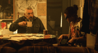 An older man with a beard drinks coffee at a table next to a young man who's been blindfolded.