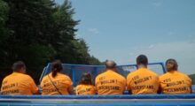 A row of people with their backs to the camera sit on a boat wearing orange t-shirts.