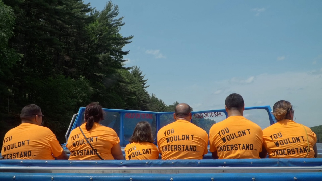 A row of people with their backs to the camera sit on a boat wearing orange t-shirts.
