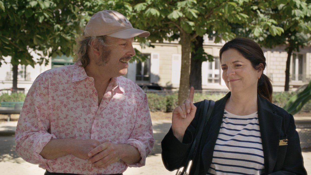 A man in a pink shirt and baseball cap stands next to a woman in a park on a sunny day.