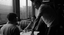 A white man leans over two people sitting at a diner booth.