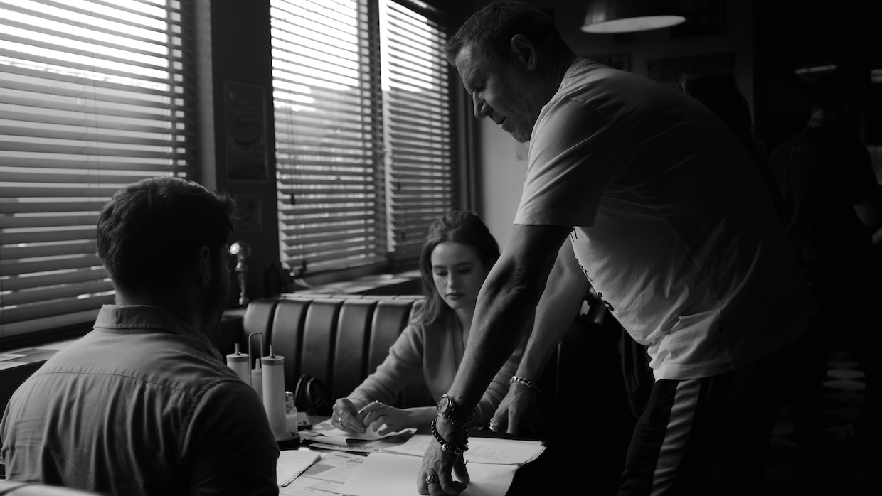 A white man leans over two people sitting at a diner booth.