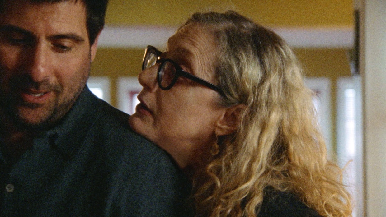 A middle-aged white woman leans her chin on a younger white man's shoulder.