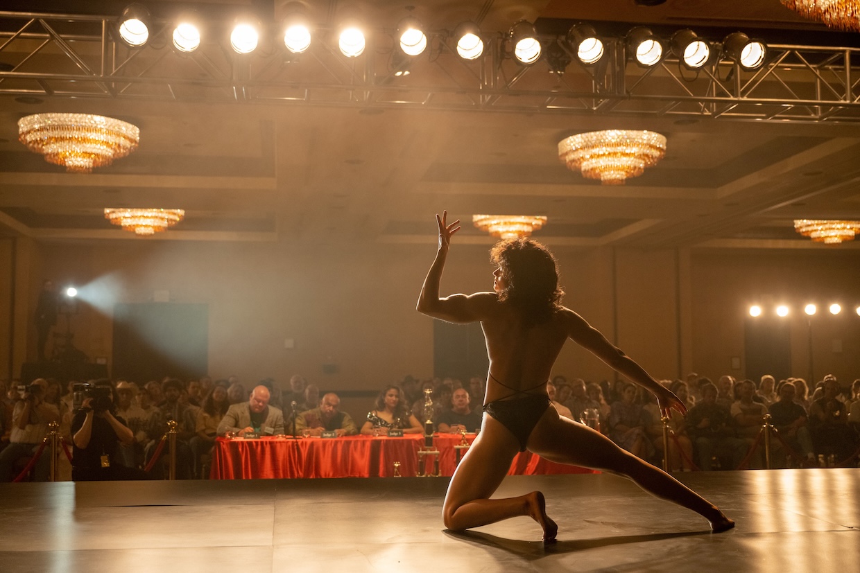 A female bodybuilder with her back to the camera flexes before a table of judges at a bodybuilding pageant.