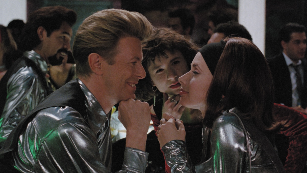 A white man and woman smile at each other across a table at a club while another woman stands between and behind them.