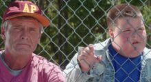 A white man and young white boy stare from behind a chain-link fence at a baseball game.