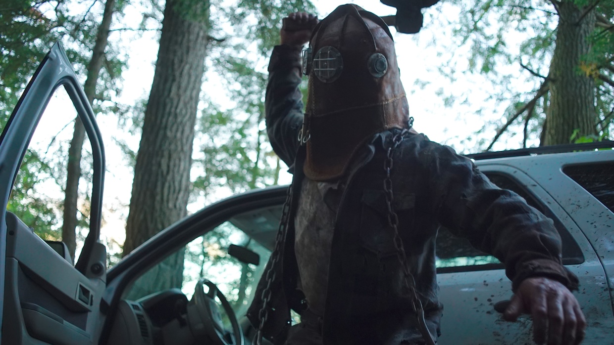A man wearing a leather helmet strikes a blow in front of a car parked in the forest.