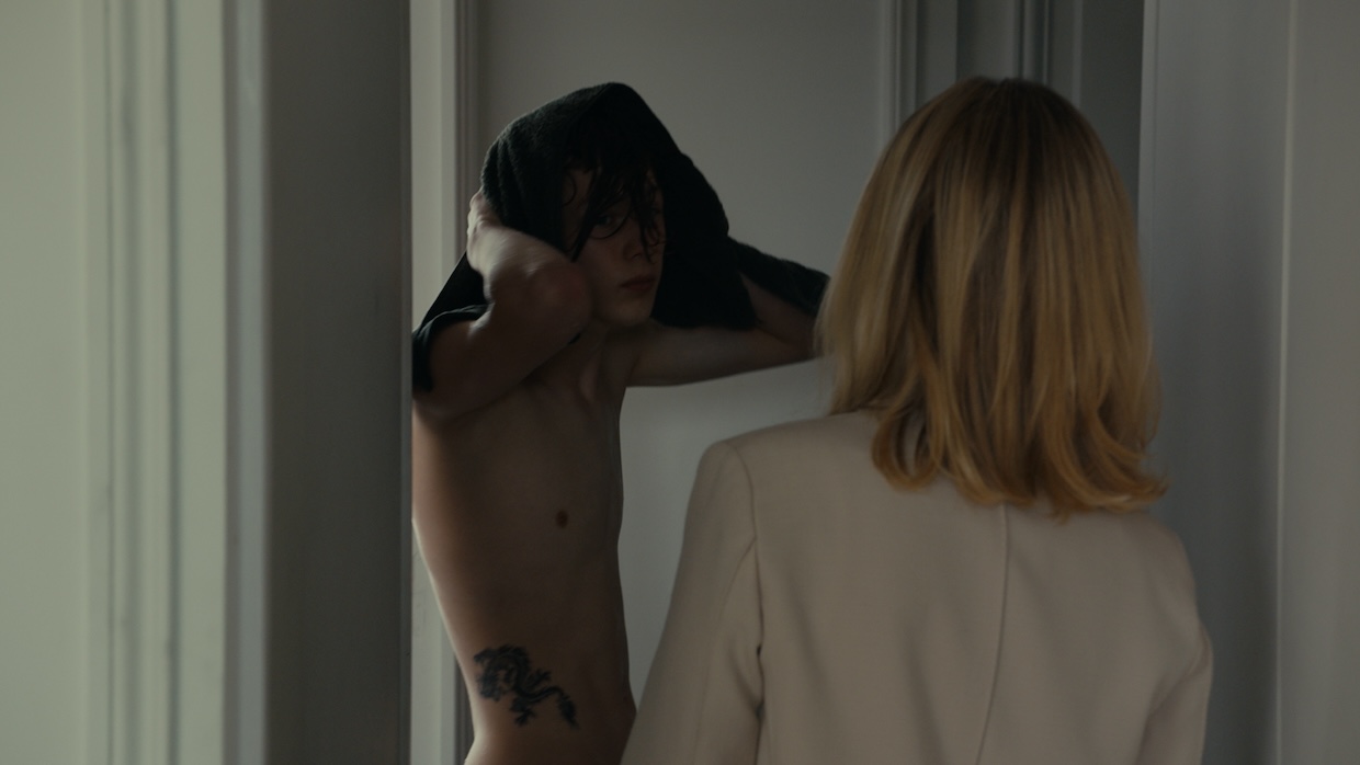 A white teenage boy pulls his t-shirt over his head in front of a woman.