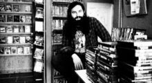 A white man with a large beard and long hair sits in a video store.