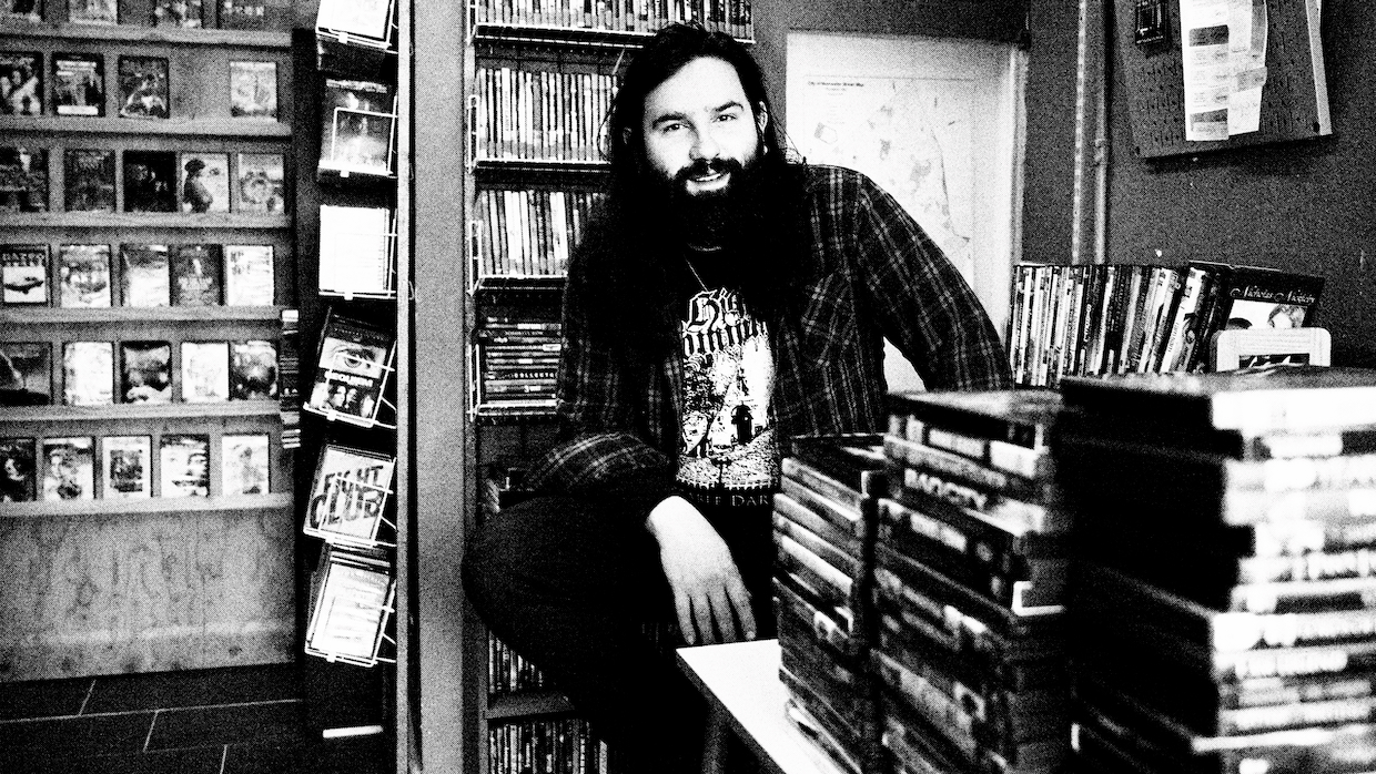 A white man with a large beard and long hair sits in a video store.