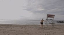 A man with his back to the camera stands facing the shoreline on a gloomy beach.