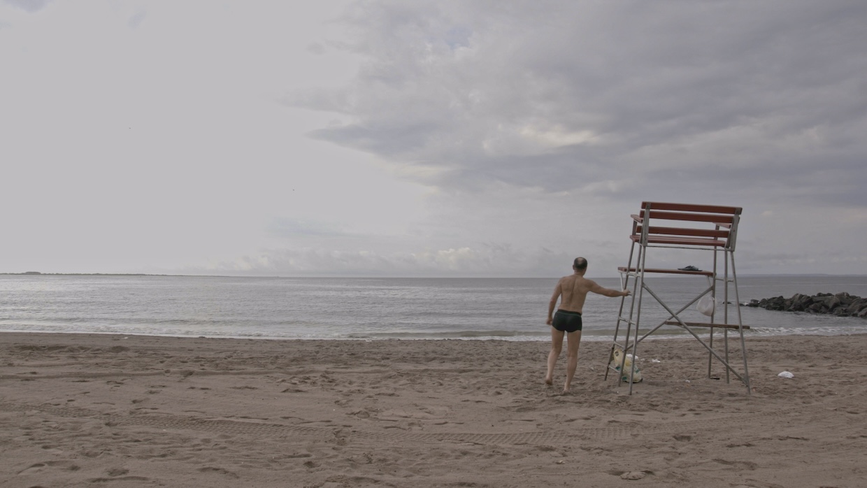 A man with his back to the camera stands facing the shoreline on a gloomy beach.