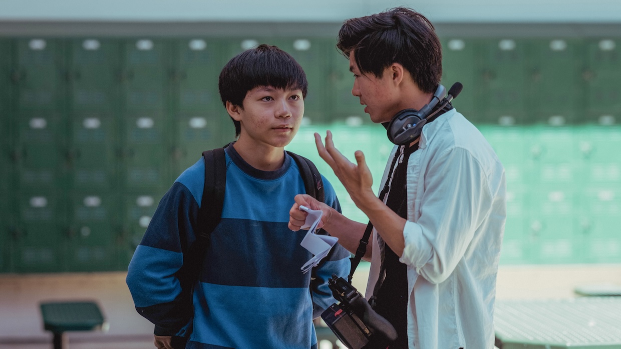 A young Asian-American teenagers listens to a 20something Asian-American director wearing headphones on a high school film set.