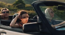 A young woman with dark sunglasses and dark hair driving in a vintage car with an older woman and man.
