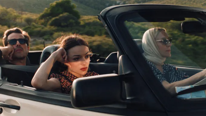A young woman with dark sunglasses and dark hair driving in a vintage car with an older woman and man.