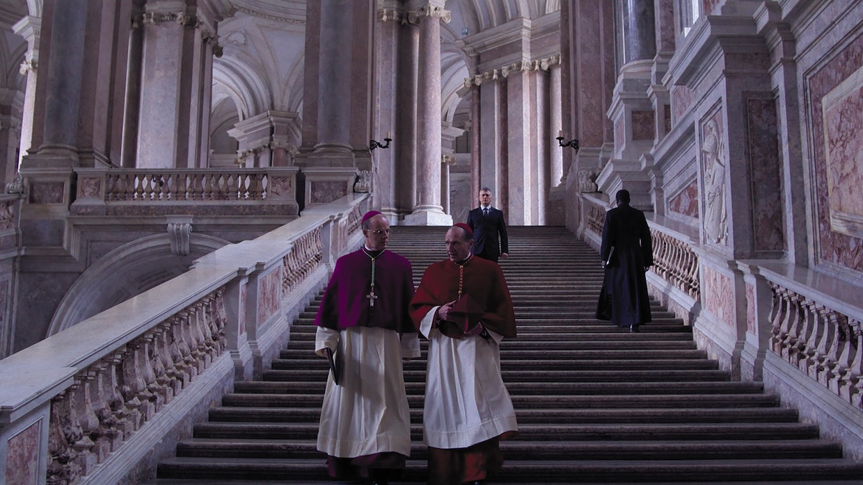 Two Catholic cardinals walk down a stairway in an elaborate building.