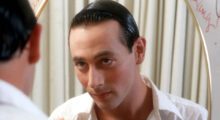 Paul Reubens looks into the mirror. He wears a white dress shirt and has his black hair slicked back.