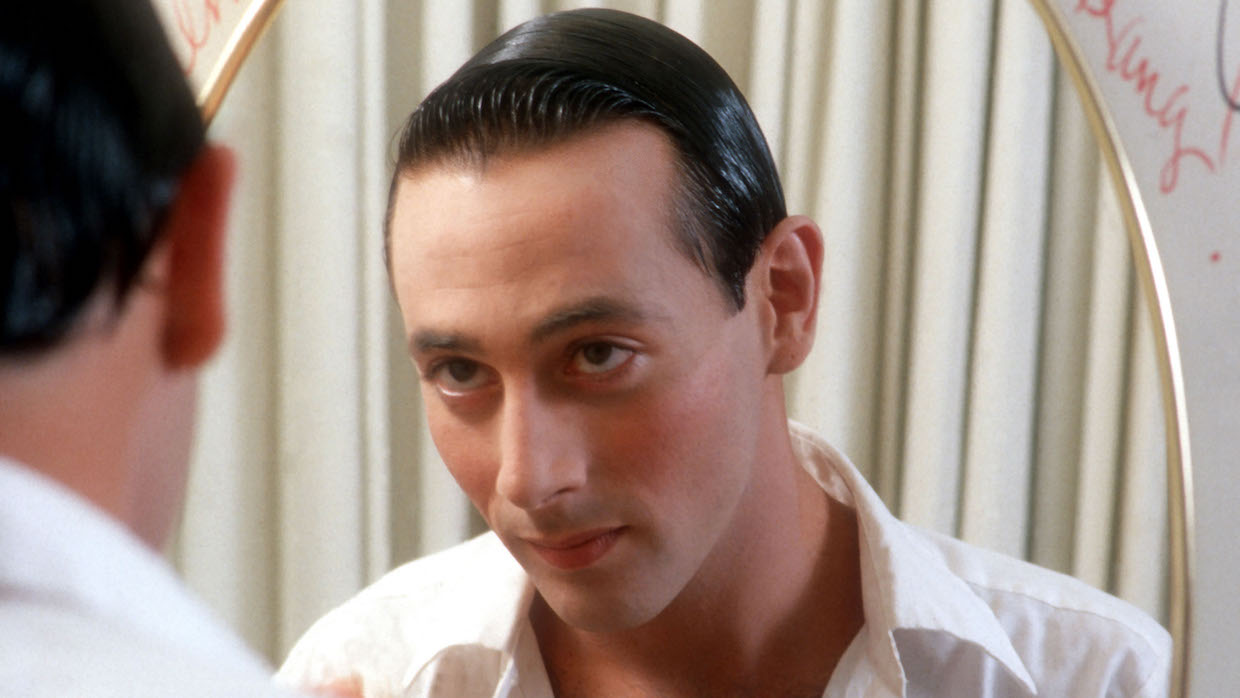 Paul Reubens looks into the mirror. He wears a white dress shirt and has his black hair slicked back.