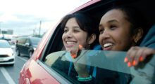 Two teenage girls of Middle East/North African descent smile while looking out the window of a red car.
