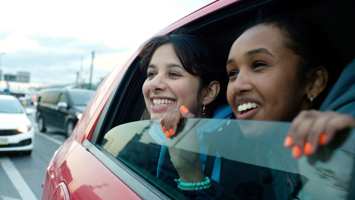 Two teenage girls of Middle East/North African descent smile while looking out the window of a red car.