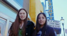 Two-shot of teenage girls on the sidewalk with a movie theater marquee in the background.
