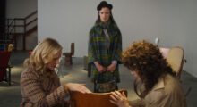 A woman in a green and blue coat watches as two other women caress a chair.