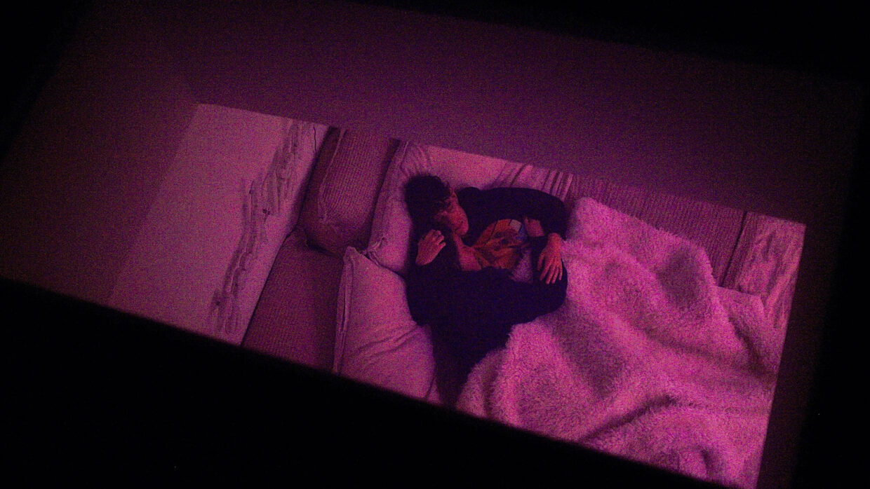 A couple bathed in a purple light cuddles on a bed.