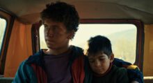Two boys, one a teenager and one younger, are sitting inside. The younger is asleep.