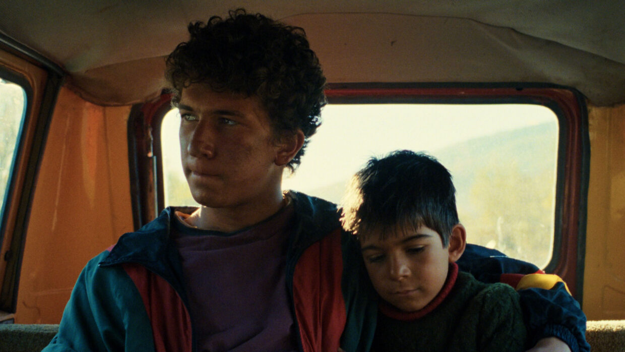 Two boys, one a teenager and one younger, are sitting inside. The younger is asleep.