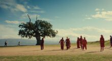 People wearing traditional garb walk through a field, a tree is off in the distance.