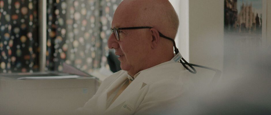 A bald man wears a doctors coat and classes.