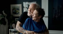 An older man with white hair and beard hugs a middle-aged woman with short black hair and a blue sweater.