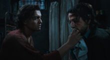 Two men are sitting on a prison floor. One has his hand near the other's mouth.