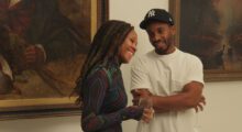 A Black couple walk through a gallery; the man wears a white t-shirt and Yankees cap, the woman wears a colorful mesh top and holds a glass of white wine.