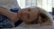 Blonde-haired actress Marlee Matlin lays down on a bed, lightly smiling and looking into the camera.