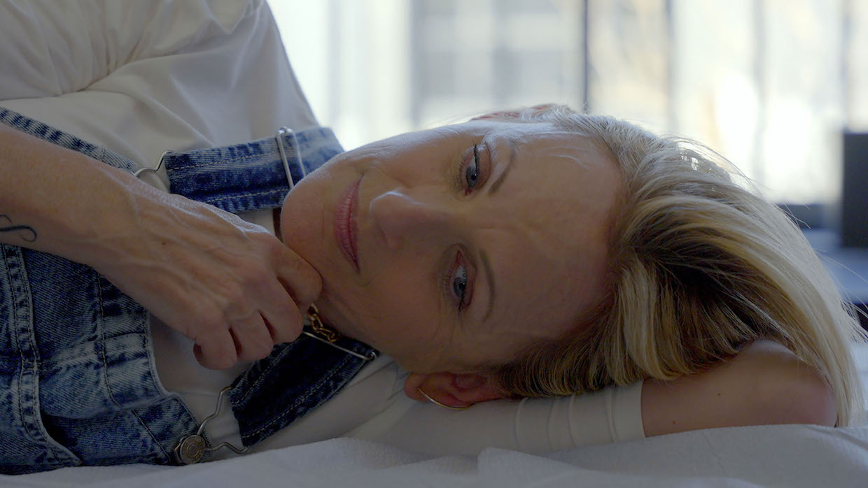 Blonde-haired actress Marlee Matlin lays down on a bed, lightly smiling and looking into the camera.