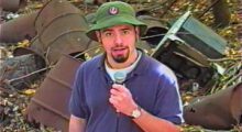 A young man wears a purple t-shirt, green bucket hat and sports a goatee. He holds a microphone and stands in front of barrels of indiscriminate waste.