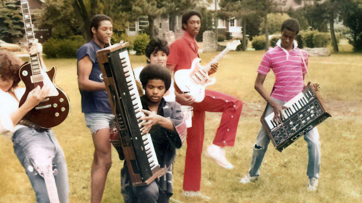 Several Black musicians are standing in the grass playing guitars and keyboards.
